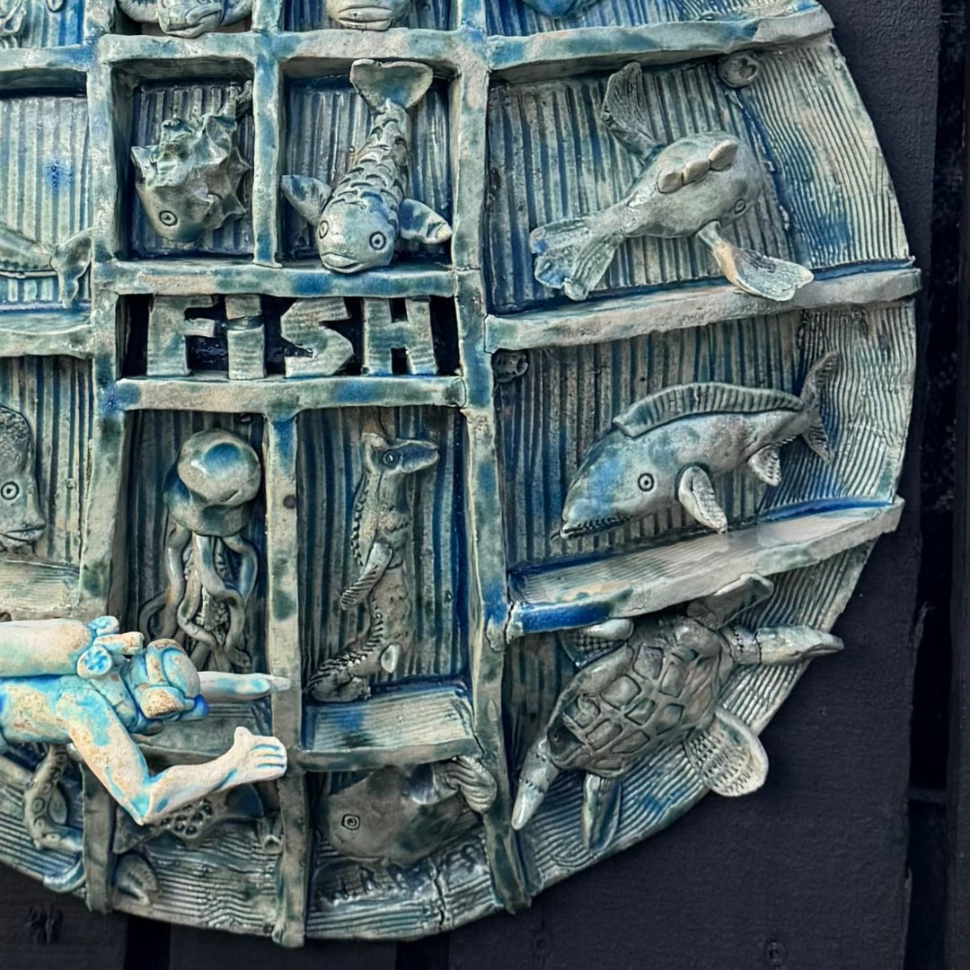 A circular textured wall plaque featuring a blue and gray relief design with various sea life illustrations, including fish, a turtle, a diver, and the word 'FISH' in the center, mounted against a dark background.