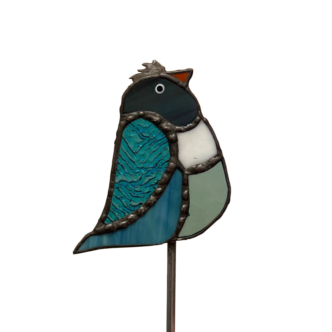 A stained glass bird figurine on a metal stake, featuring blue, teal, black, white, and sage green sections outlined in dark metal, set against a white background.