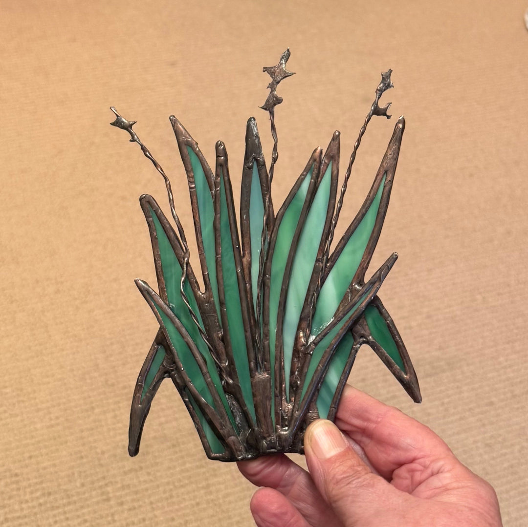 A hand holding a stained glass art piece depicting green agaves or ornamental grass with dark metal leading, set against a neutral tan background.