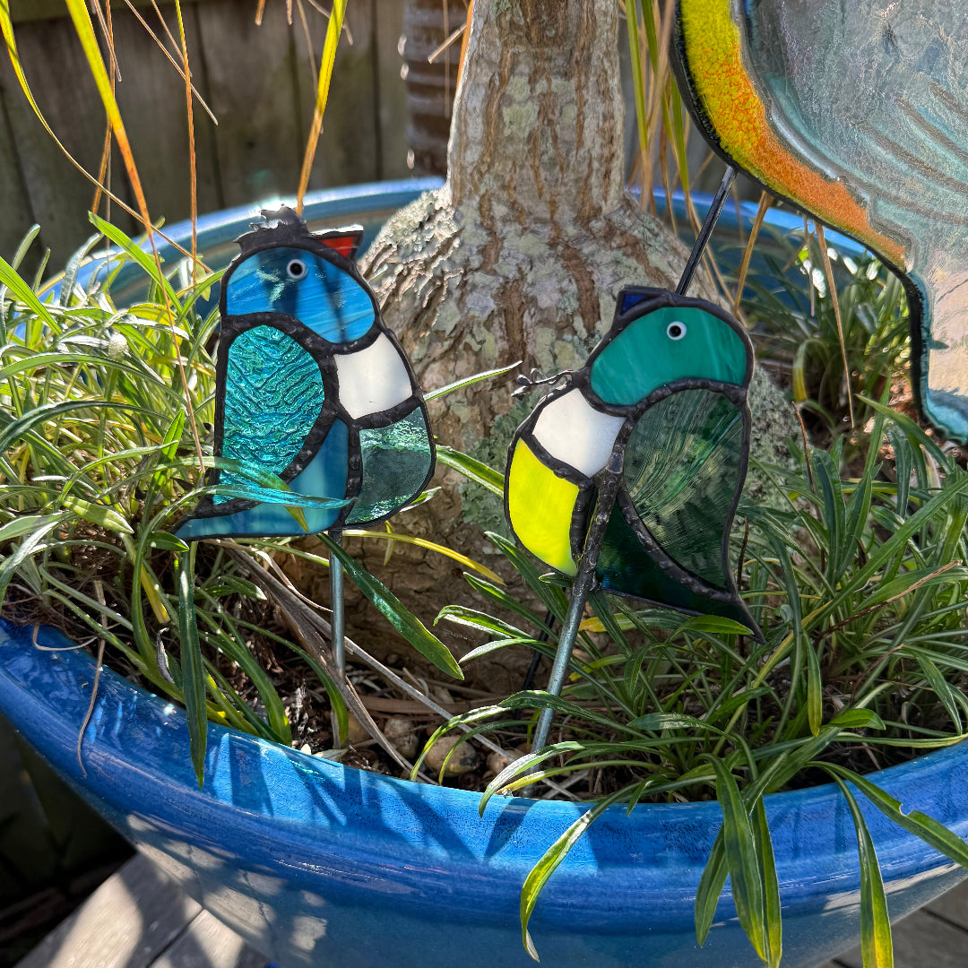 Two colorful stained glass bird garden stakes placed in green grass inside a blue planter pot outdoors.