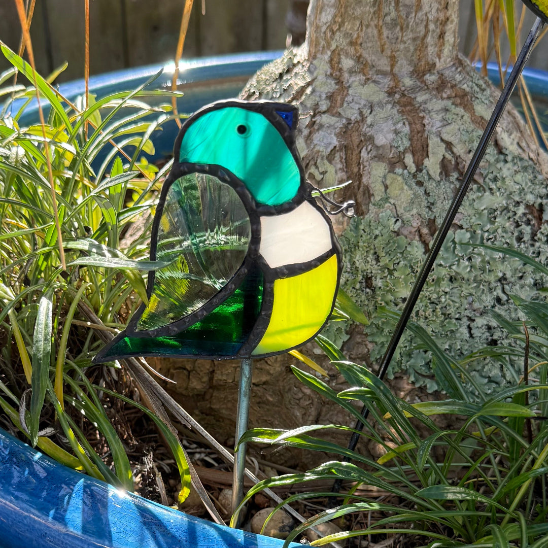 A stained glass bird garden stake with a teal, white, and yellow design, displayed in a garden next to a tree trunk and plants.