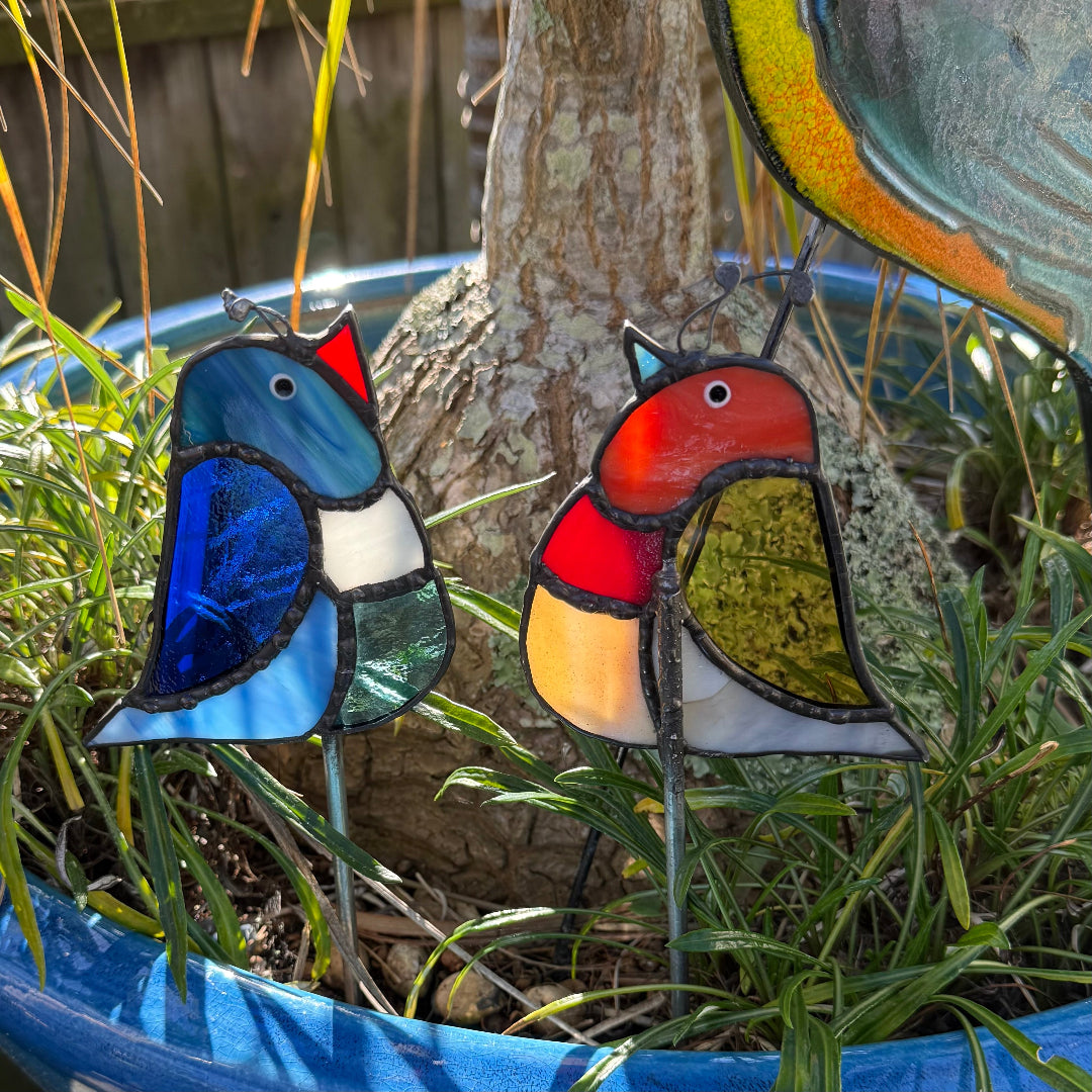 Two stained glass bird garden stakes with colorful, multi-sectioned bird designs placed in a garden planter next to a tree trunk.