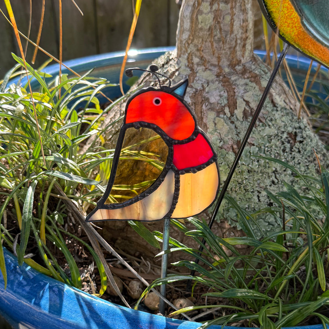 A stained glass bird garden stake with a colorful bird design, placed in a blue planter among green grass and a tree trunk.