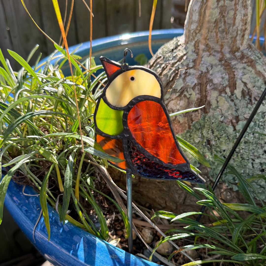 A stained glass bird garden stake with a colorful bird design, placed in green grass next to a tree trunk in a blue planter.