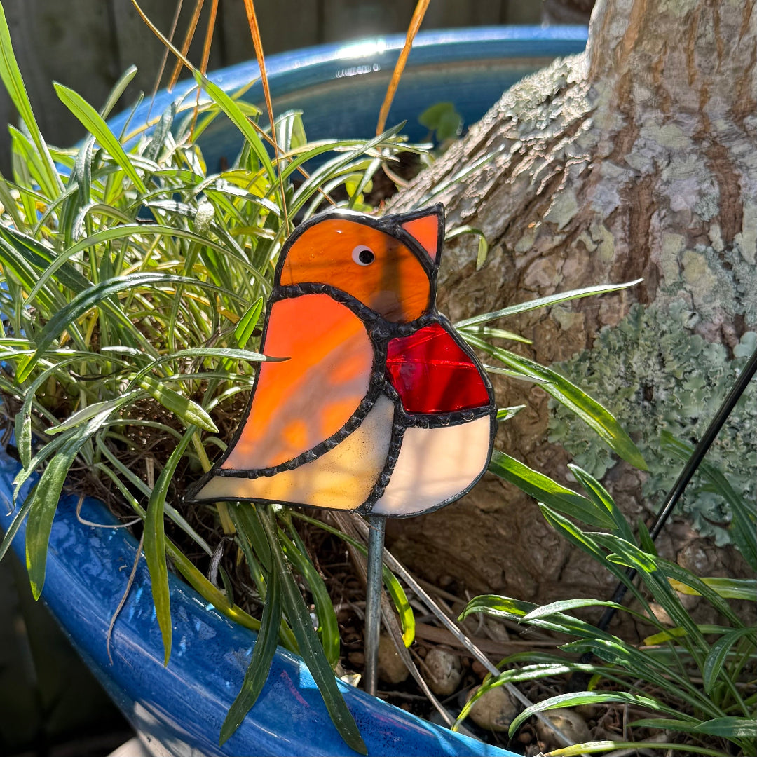 A stained glass garden stake shaped like a robin bird, featuring orange, red, and cream sections outlined in black, displayed in a blue planter among green grass and a tree trunk.