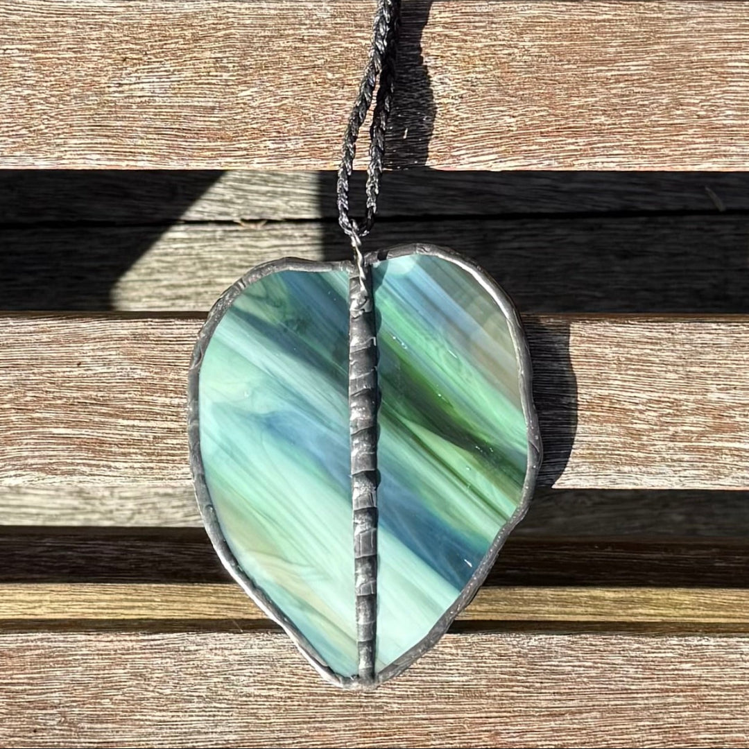 A Kawakawa Leaf made of opaque iglass with green, blue, and cream tones, suspended on a dark cord over a weathered wooden slat background.