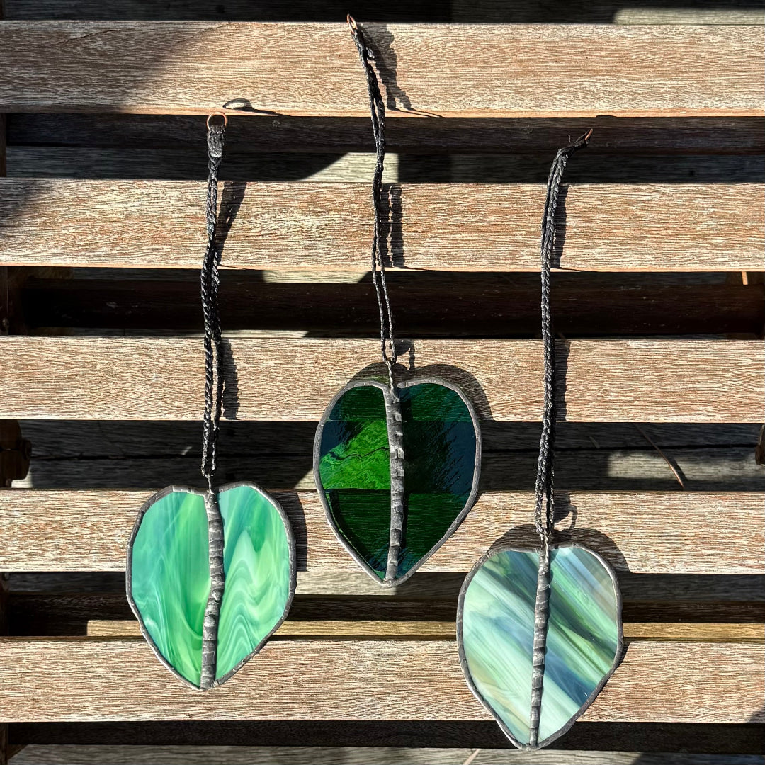 Three stained glass leaf suncatchers in shades of green and white, hanging on a rustic wooden slat wall.
