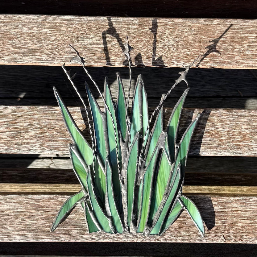 A green succulent-style metal wall art piece mounted on a weathered wooden slat surface, with shadows cast above it.