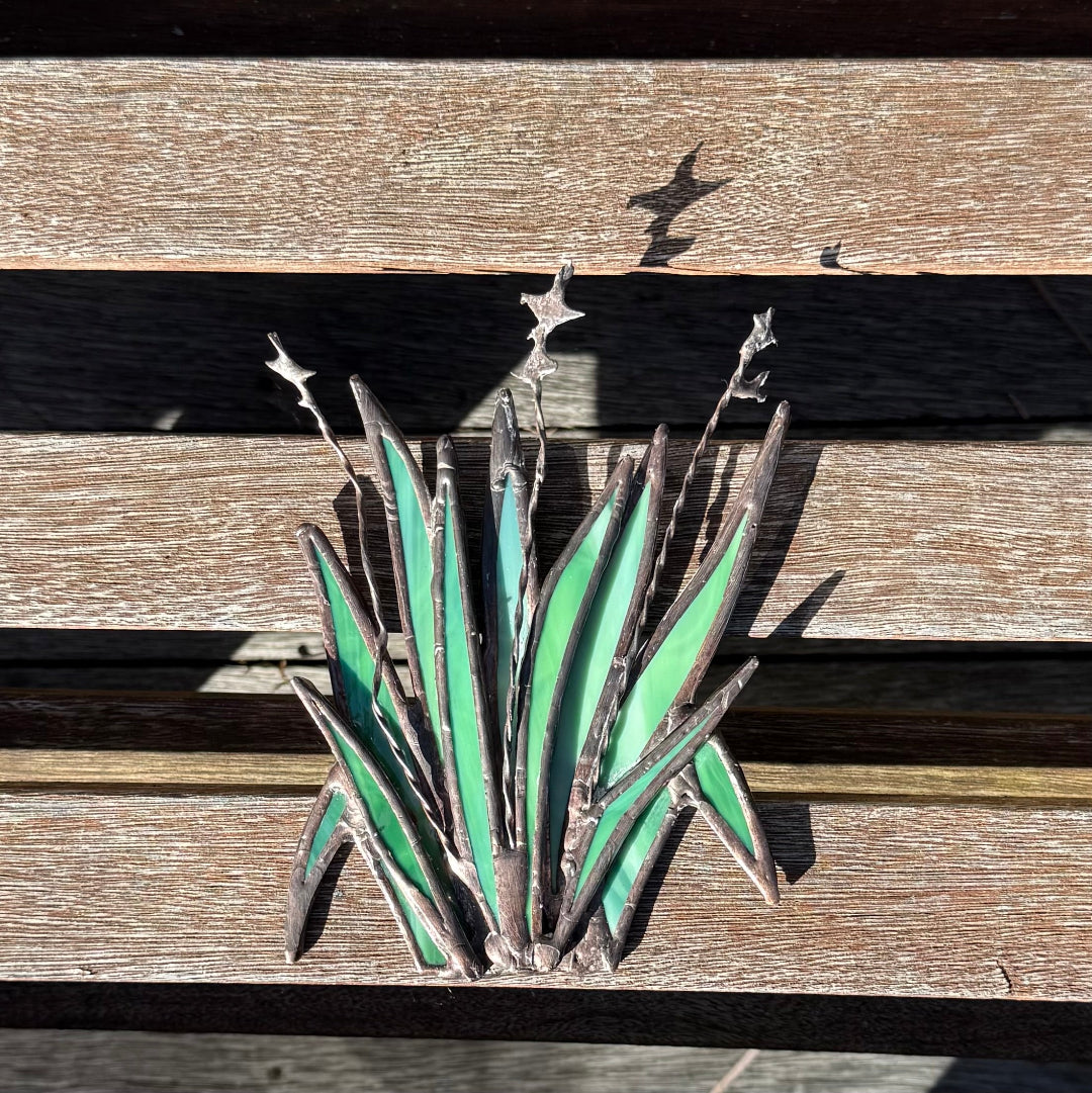 A metal garden sculpture of succulent leaves with green enamel fillings, mounted on a weathered wooden slat bench in direct sunlight.
