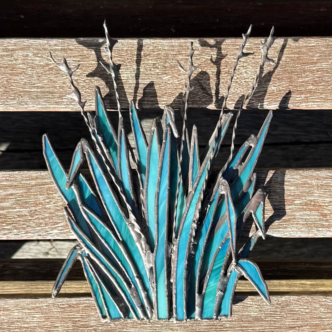 A metal wall art sculpture depicting a cluster of blue agave leaves and flowering stems, mounted on a rustic wooden plank surface.