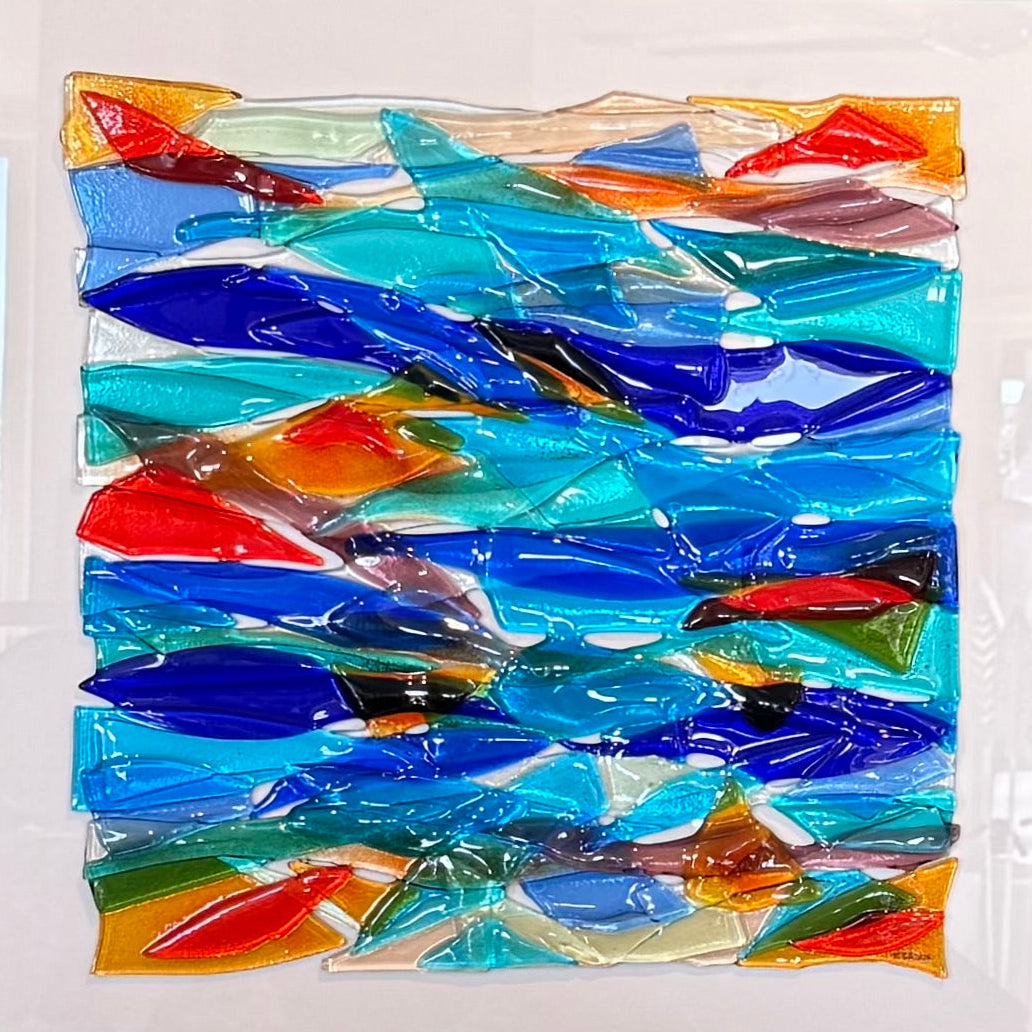 A square abstract glass mosaic art piece in a white frame, featuring layered translucent blue, teal, orange, and red glass shapes on a light gray wall.