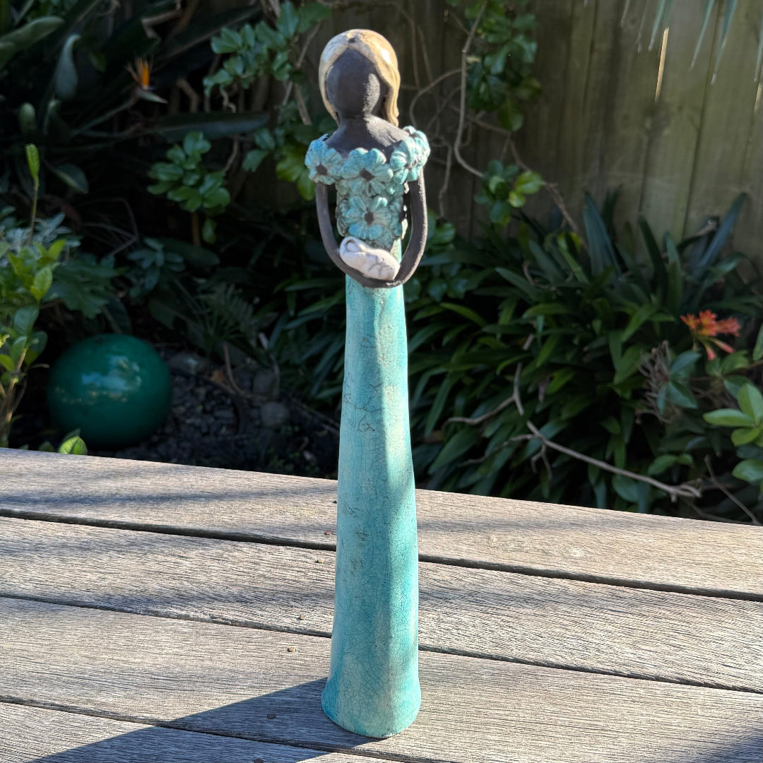 A decorative garden figurine of a woman with blonde hair, wearing a long teal dress and holding a white flower, standing on a weathered wooden table with a garden background.