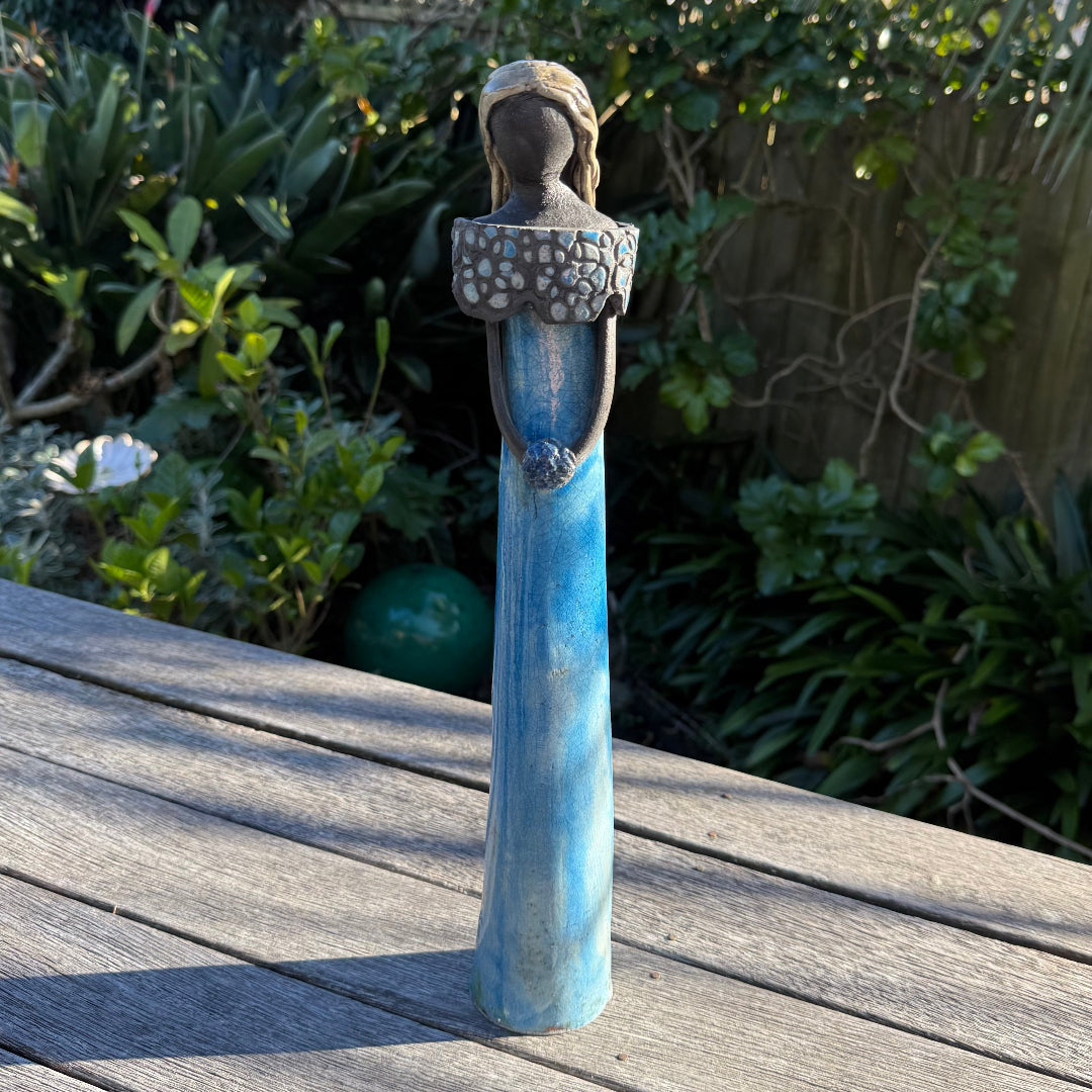 A decorative figurine of a woman with a long blue skirt and patterned top, standing on a wooden deck with a garden background.