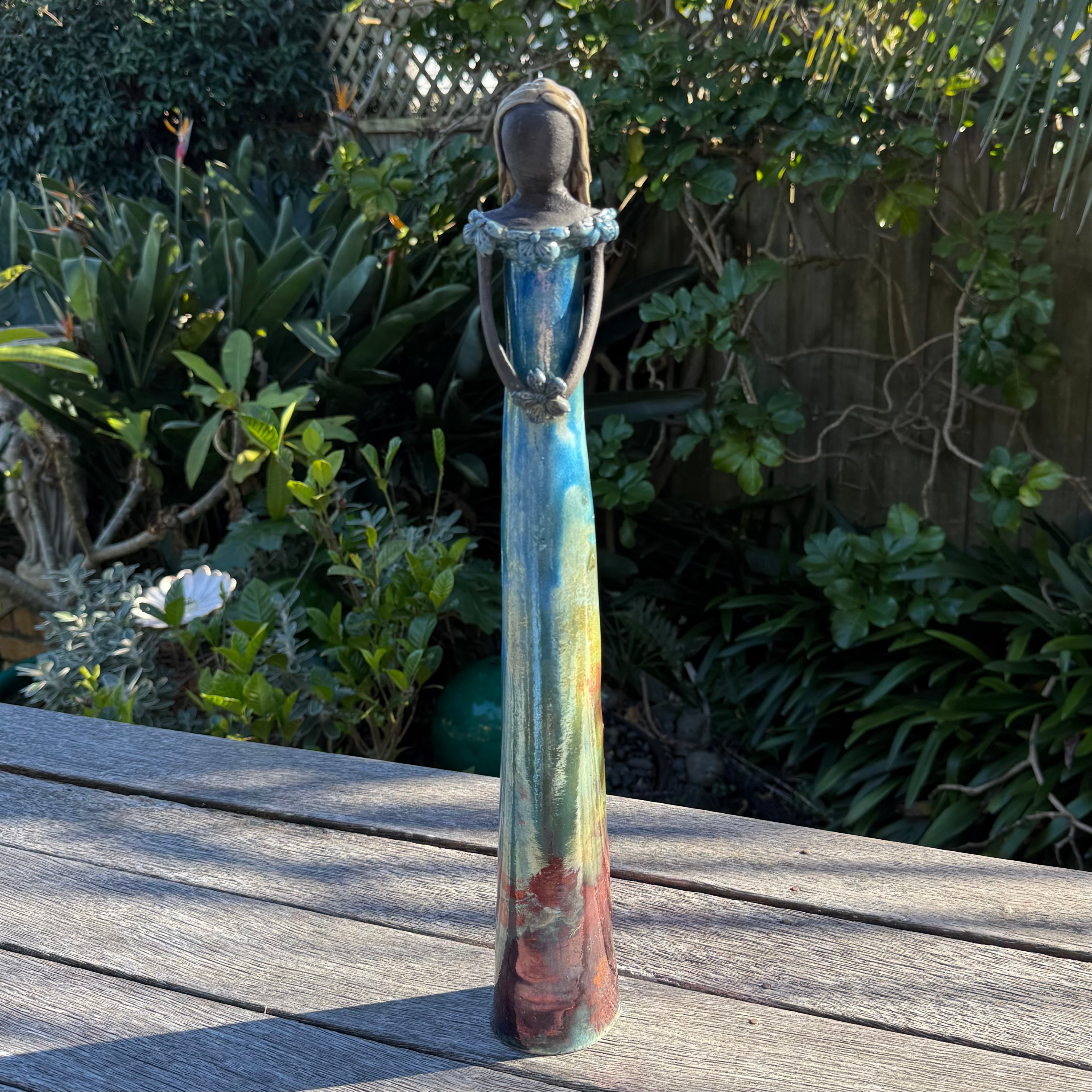 A tall, slender statue of a woman in a flowing dress, standing on a wooden deck with a lush green garden background.