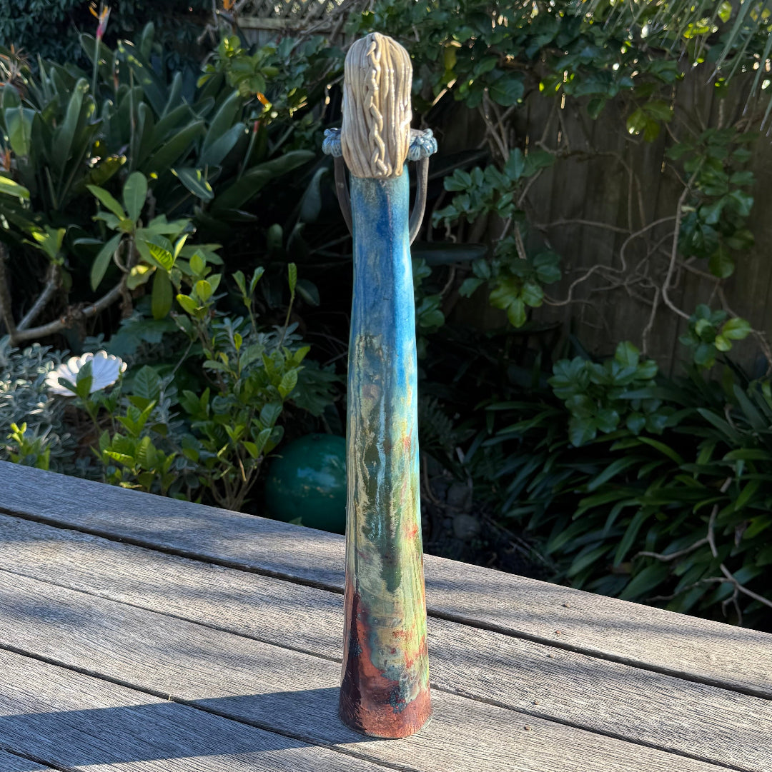 Back view of a tall, slender ceramic woman with long blonde hair and a flowing blue, green, and rust-colored robe, standing on a weathered wooden deck with a lush garden background.