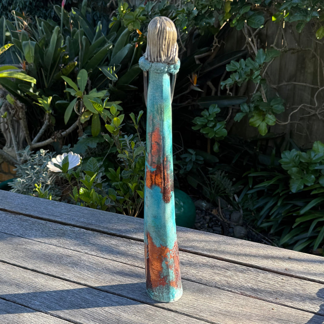 Back view of a tall, slender decorative ceramic figurine of a woman with long dark hair, wearing a turquoise and rust-colored dress, standing on a weathered wooden deck with a lush garden background.