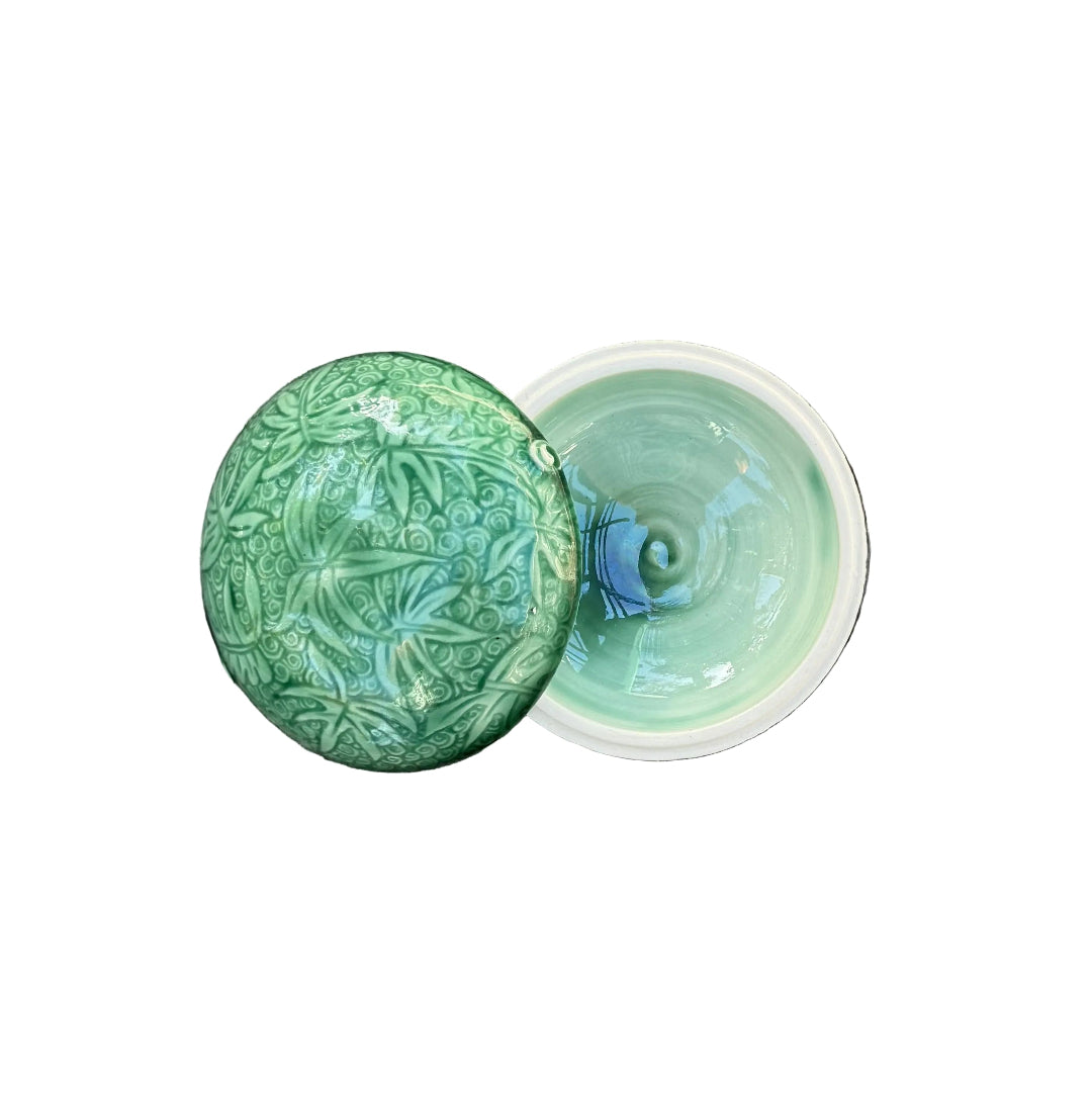 A small round ceramic lidded container with a green embossed floral lid and a pale mint-green interior, placed on white  surface.