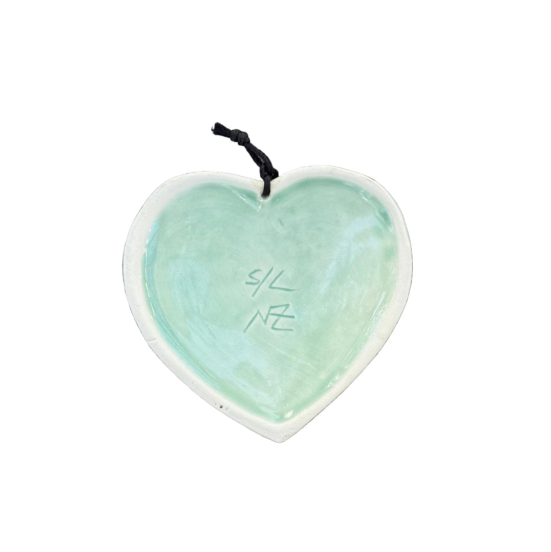 Back view of a heart-shaped ceramic wall hanging ornament with a white rim and a pale green glazed center, signed S/L NZ.