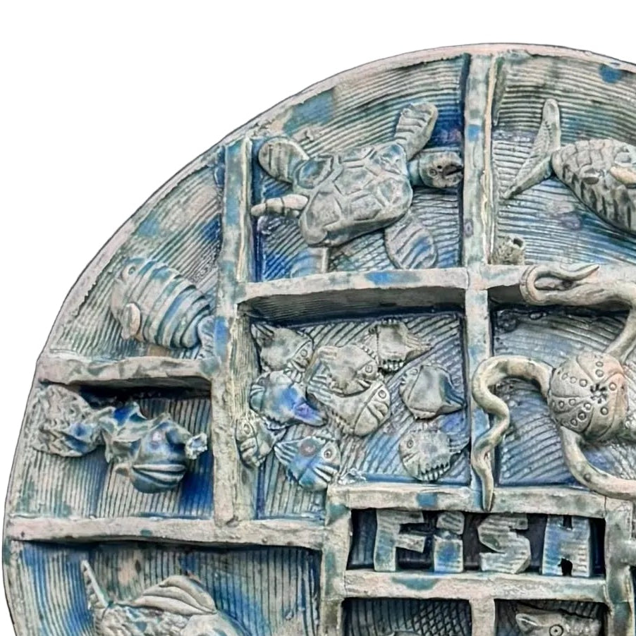 Close up view of a round, multi-compartment ceramic wall sculpture featuring relief carvings of various sea creatures, including fish, a turtle, an octopus, a seahorse, and a shark, with a blue and white shark mounted in front on the lower left, set against a white background .