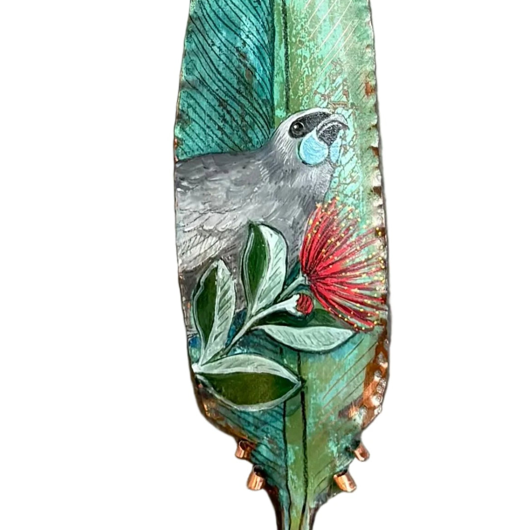 Close up view of a copper feather wall art sculpture hand painted with a Kokako bird on Pohutakawa flowers against a white background.