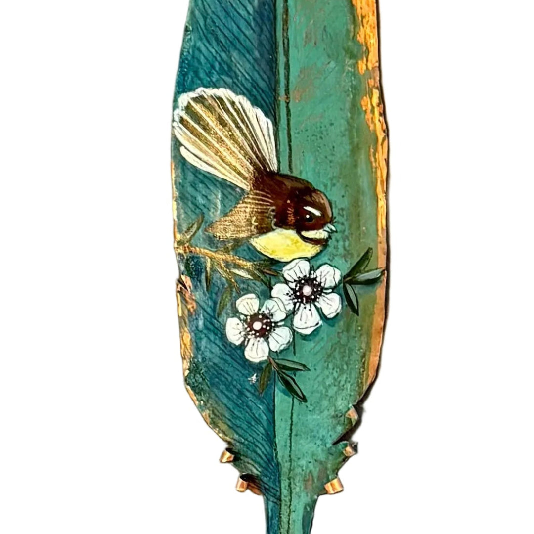 Close up view of a copper feather wall art sculpture hand painted with a Fantail bird on Manuka flowers against a white background.
