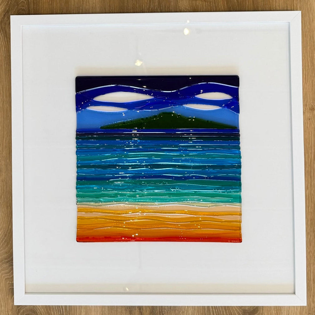 A square framed artwork on a light wood background, featuring an abstract seascape made of glass with dark blue sky, layered blue and teal water, and a sandy shore in orange, yellow, and green.