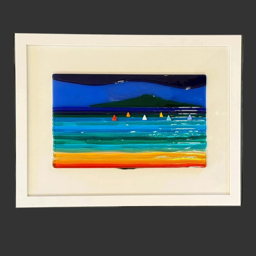 A framed glass seascape of a coastal scene with deep blue sky, turquoise water, green hills, and an orange shoreline, mounted on a gray wall.