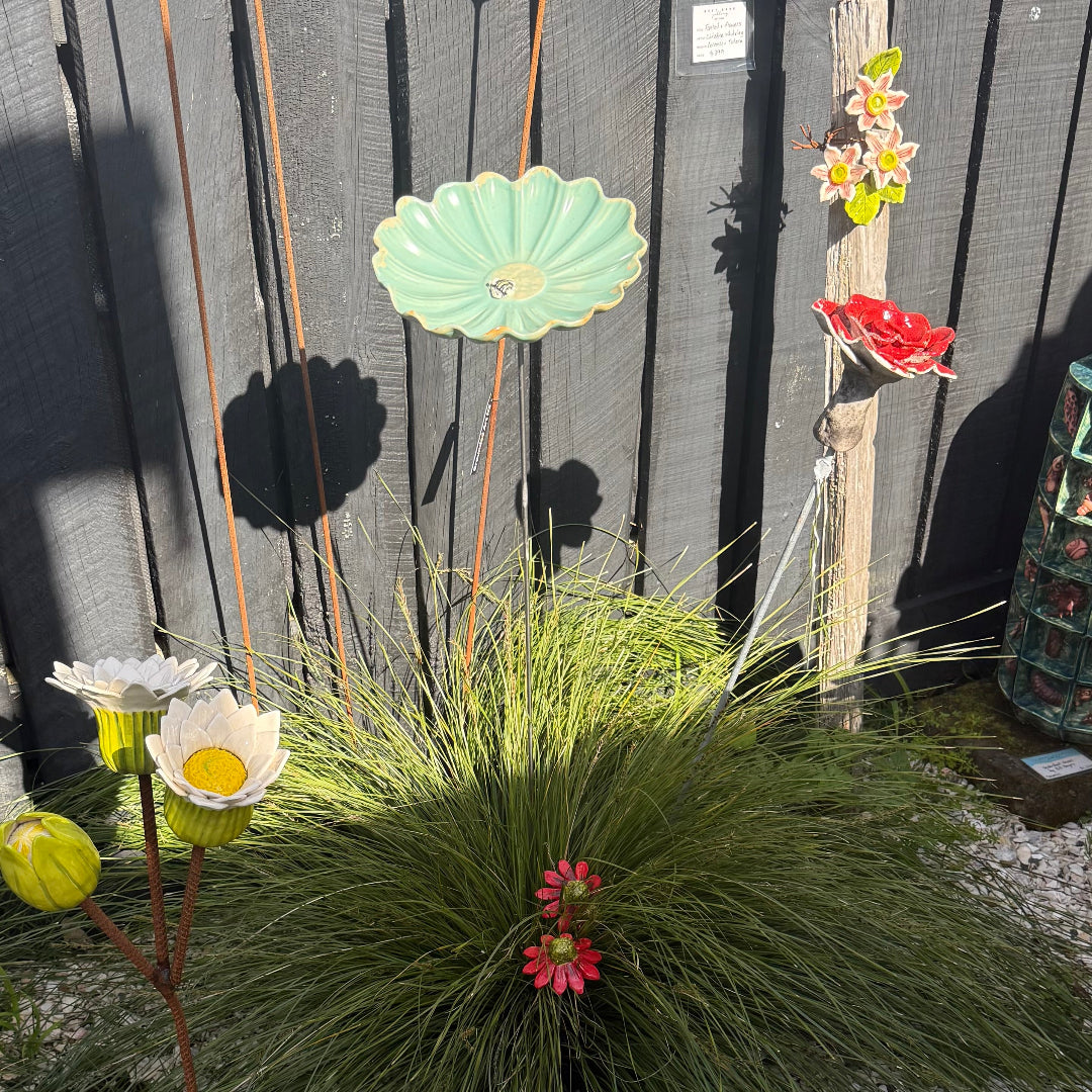 A cluster of decorative garden metal flower sculptures in various colors and styles, including a mint green daisy with a bee, white daisies, and red blossoms, arranged in front of a dark gray wooden fence.