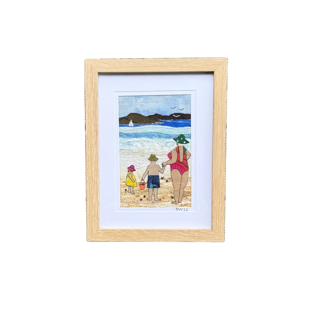 A small framed beach textile artwork with a light wood frame and white mat, placed on a white background. The artwork depicts a beach scene with a woman & 2 children playing.
