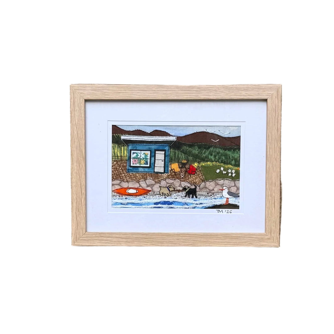 A small framed textile landscape artwork depicting a blue bach on a white background.