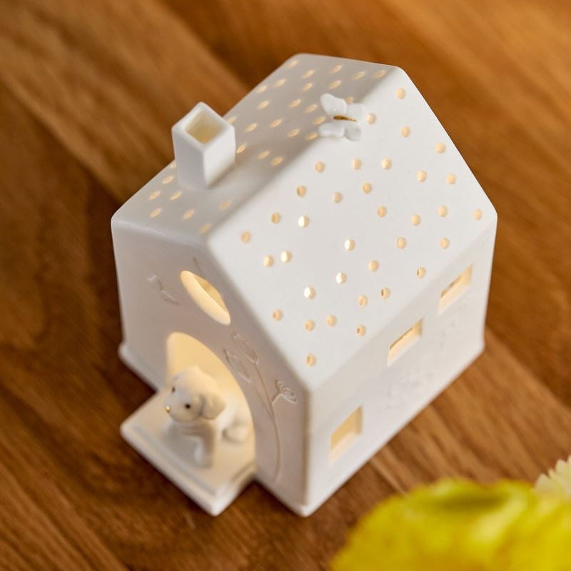 A white ceramic house-shaped tea light holder with a small dog figure on the porch, shown lit on a wooden surface with blurred yellow flowers in the foreground.