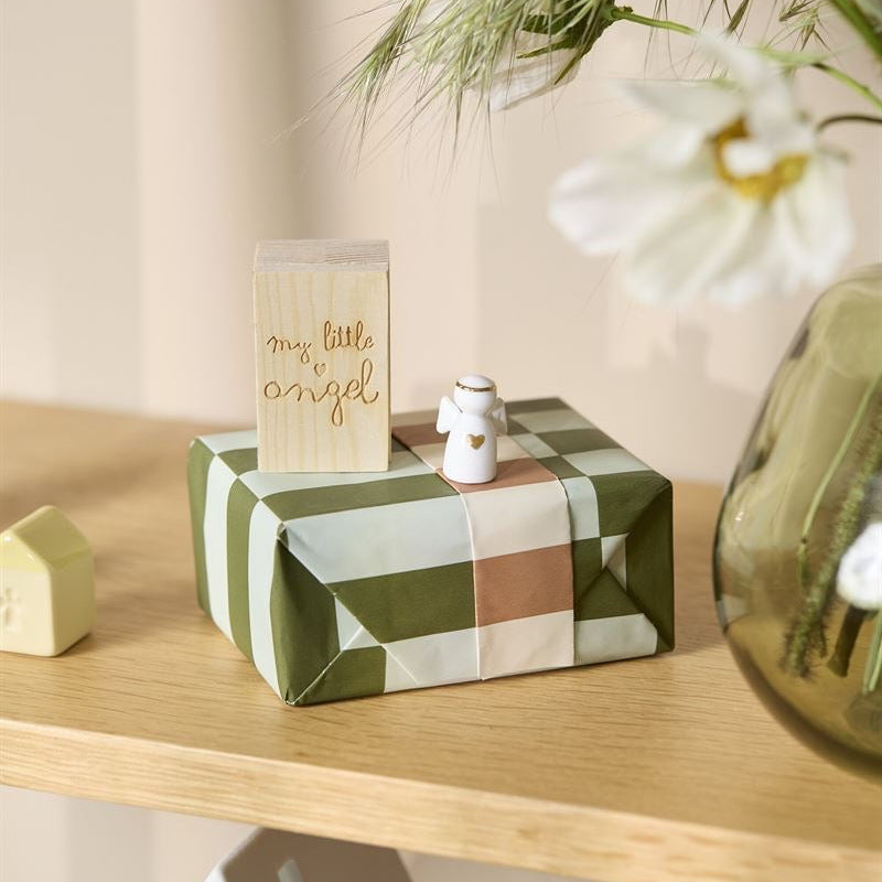 A wrapped gift box with green, beige, and white plaid wrapping paper sits on a light wood shelf, decorated with a small wooden card that reads 'my little angel' and a tiny white snowman figurine. A vase with white flowers is blurred in the foreground.