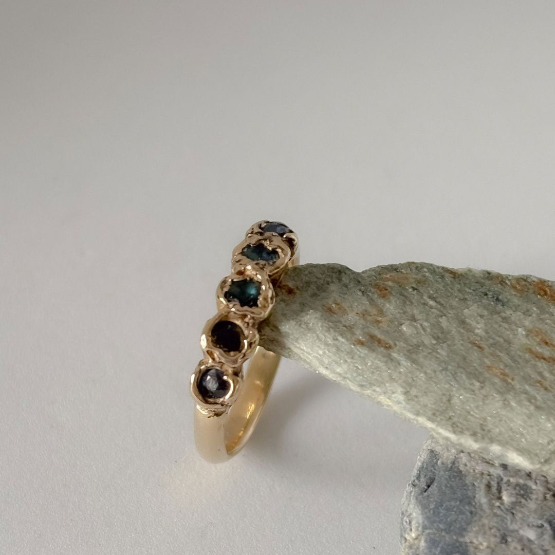 A gold ring with five blue gemstones, displayed on a piece of textured gray stone against a neutral light gray background.