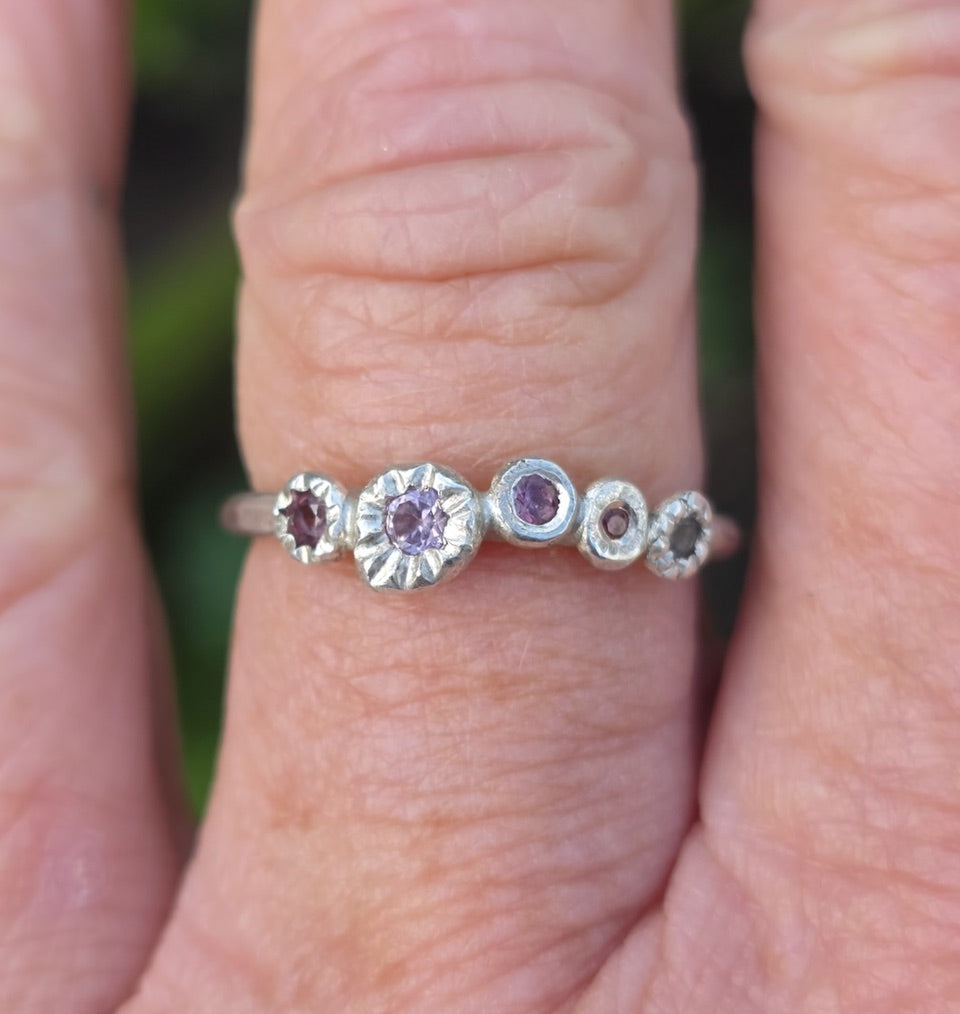 A close-up of a hand wearing a silver ring with five small floral clusters set with purple gemstones.