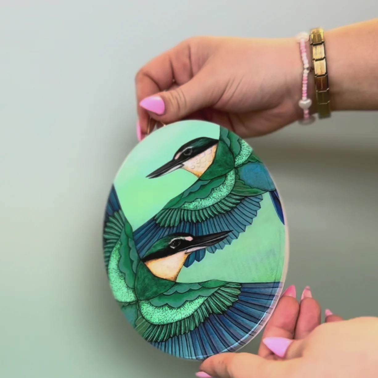 Oval wall tile with a green and blue bird design held by a person against a light gray background