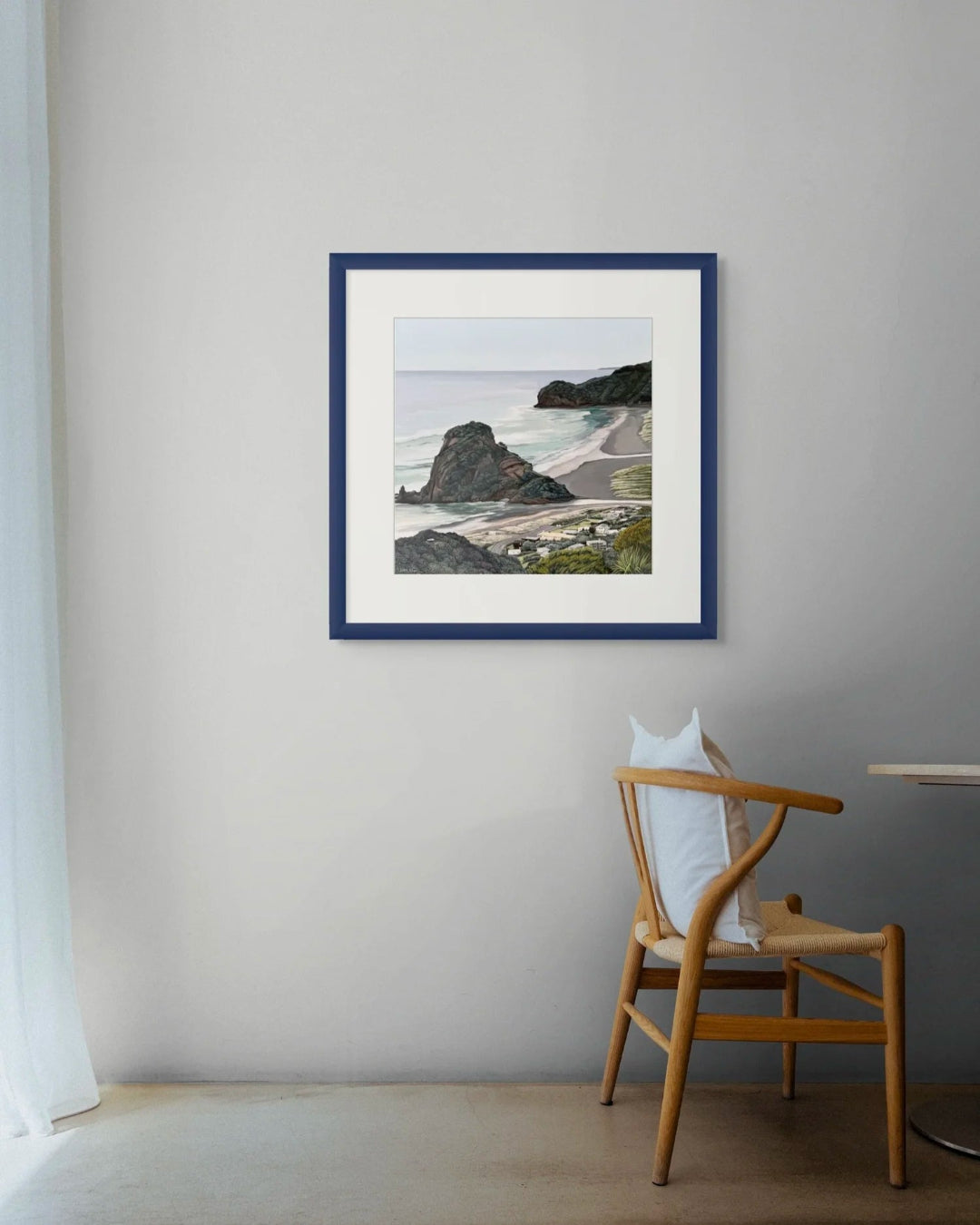 A framed coastal landscape print hangs on a light gray wall above a wooden chair in a minimalist room setting.