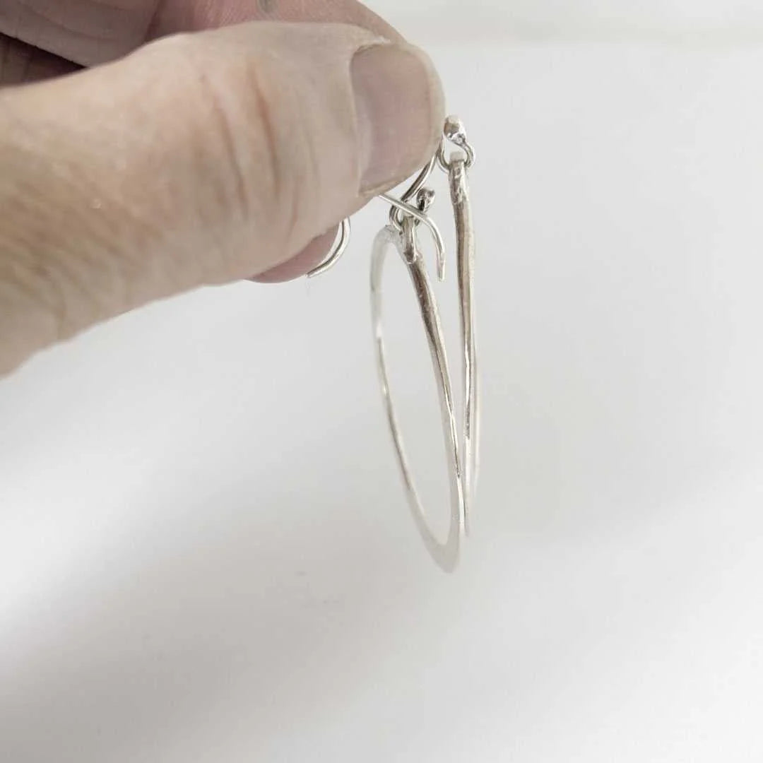 A close-up of a hand holding a pair of thin silver hoop earrings against a plain white background.
