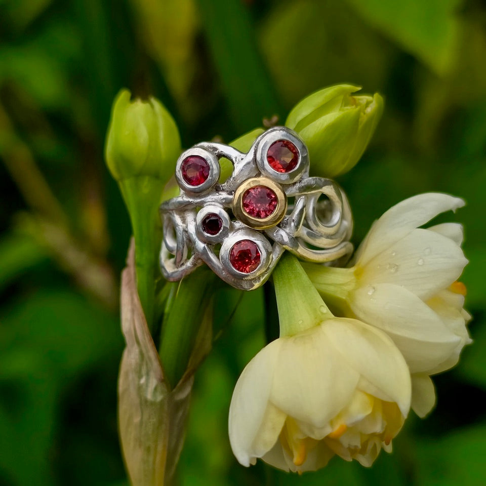 A silver ring with orange gemstones and a gold-toned center setting, displayed on a green flower bud and pale cream bloom against a soft green background.