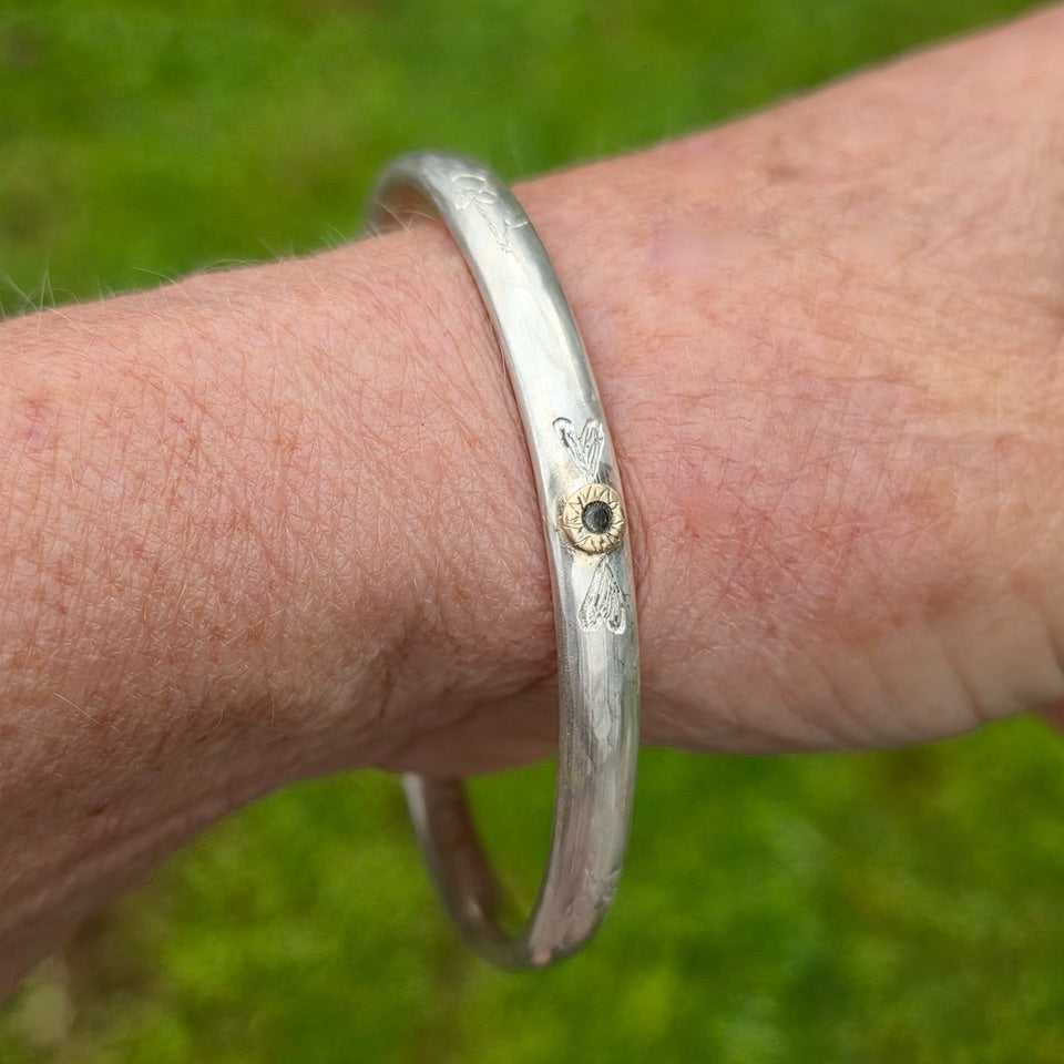 A sterling silver bangle with engraved floral details and a small green sapphire set in engraved gold, shown worn on a person's wrist against a blurred green grass background.