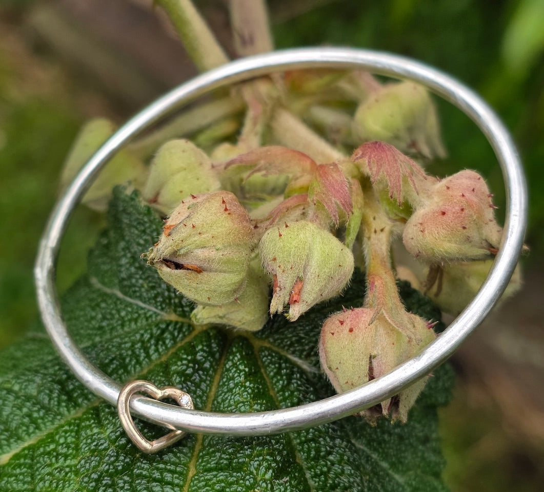 A sterling silver bangle with a small gold heart with diamond, shown resting on a green leaf with small pinkish-green berries under natural light.